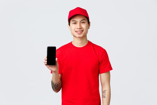 Delivery application, online shopping and shipping concept. Smiling delivery guy in red uniform, cap and t-shirt, showing mobile phone screen, advice download app for bonus to order.