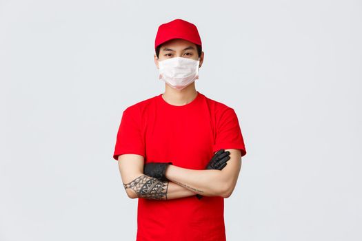 Confident young asian man in red delivery service uniform and cap, wearing protective medical mask and gloves ready to transfer your goods. Safe shopping concept. Courier cross arms on chest.