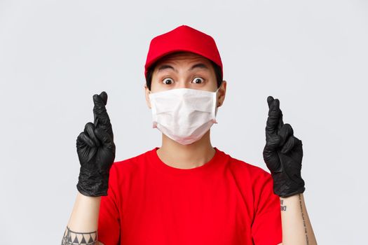 Covid-19, self-quarantine online shopping and delivery concept. Nervous asian courier in medical mask and protective gloves, wear uniform, cross fingers praying, hoping something fulfill.