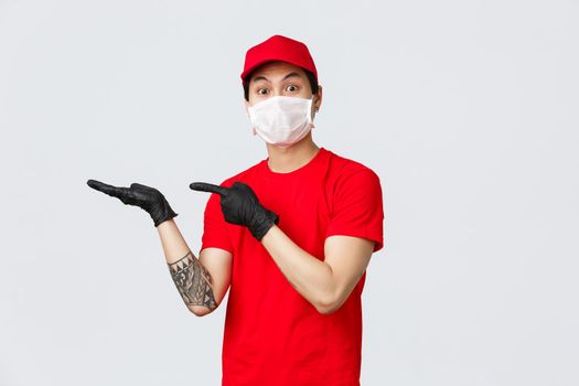 Look at this. Excited asian delivery man in red cap, t-shirt, wearing protective latex gloves and medical mask, showing something in hand, pointing at palm and stare camera impressed.
