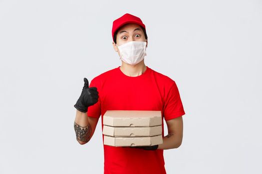 Funny charismatic asian delivery man in red uniform, cap and t-shirt. Wearing protective gloves, medical mask for clients safety during covid-19 delivery. Holding pizza boxes and make thumb-up.