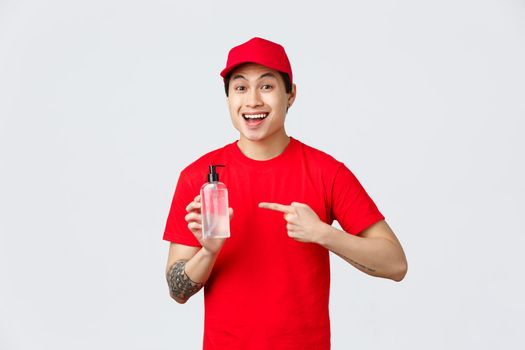 Quarantine, safe contactless delivery and shopping concept. Cheerful smiling asian courier in red cap and t-shirt, pointing hand sanitizer, promote safe shopping, importance of prevent virus spread.