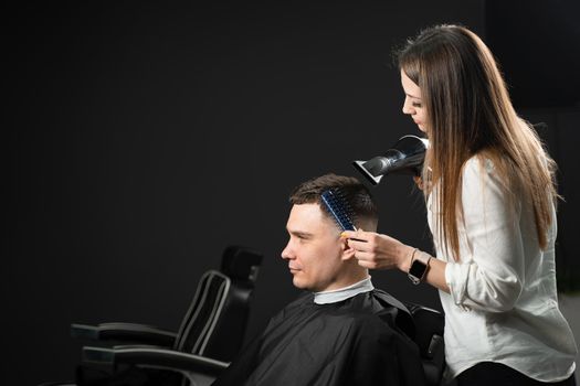 Drying hair after cutting in barbershop for handsome man. Woman hairdresser making hair style