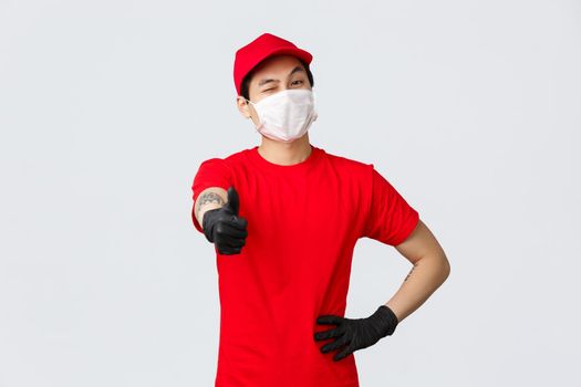 Best delivery service. Enthusiastic asian courier worker in medical mask and gloves, red uniform, show thumbs-up and wink as assuring best quality of delivery service, no problem, deliver in time.