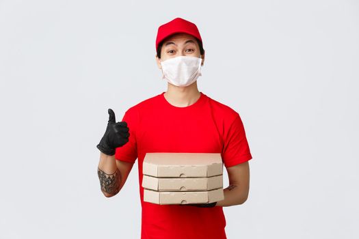 Friendly young delivery guy bring pizza to your doorstep during self-isolation quarantine on covid 19 outbreak, wearing medical mask and gloves customers safety, thumb-up, smiling.