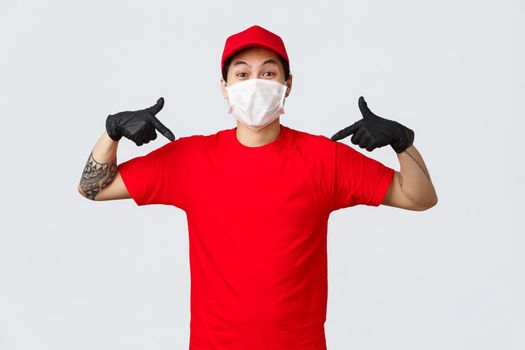 Happy asian delivery guy in red uniform, cap and t-shirt, wearing protective gloves and mask during covid19 quarantine, pandemic. Pointing himself, make orders using safe courier services.
