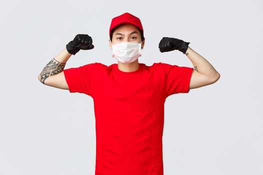 Strong confident asian delivery guy show-off, flex biceps, wearing medical mask and protective gloves. Advertisement of safe and fast courier service during pandemic covid 19 self-isolation.
