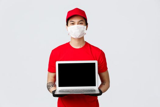 Place your order and we deliver fast. Smiling asian delivery guy showing online shopping site for clients during pandemic covid-19 wear protective medical mask and gloves, holding laptop.