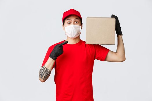 Concept of delivery and carriers during coronavirus pandemic. Wonder what inside. Curious asian courier holding box on shoulder and pointing at package, wear protective gloves and medical mask.