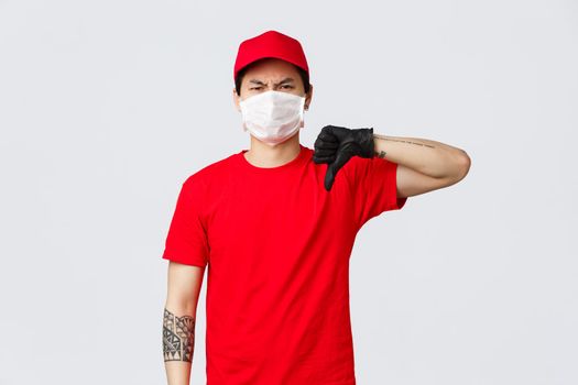 Another deliveries awful. Reluctant, disappointed delivery man in red cap, t-shirt, wearing protective gloves and medical mask, grimacing dislike and disagree, show thumb-down negative bad opinion.
