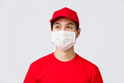 Covid-19, self-quarantine online shopping and delivery concept. Thoughtful young asian courier working during coronavirus outbreak to bring orders client doorstem, wear medical mask and uniform.
