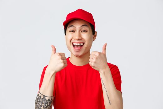 Delivery, contactless orders and shopping concept. Supportive cheerful young asian courier in red uniform cap and t-shirt, show thumbs-up in approval, recommend deliver service, grey background.