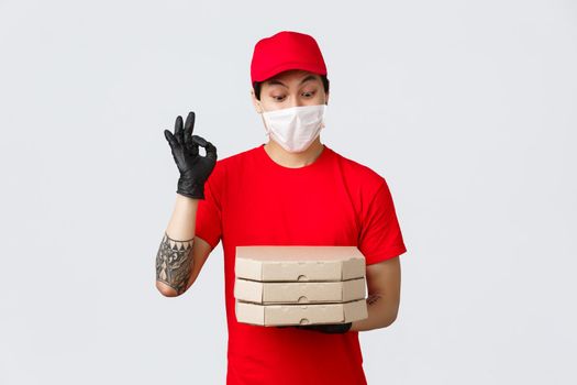 Amused asian delivery man red uniform, cap and t-shirt, wearing protective gloves and medical mask for client safety as deliver food to doorstep, bring pizza, show okay sign and stare at tasty pizza.