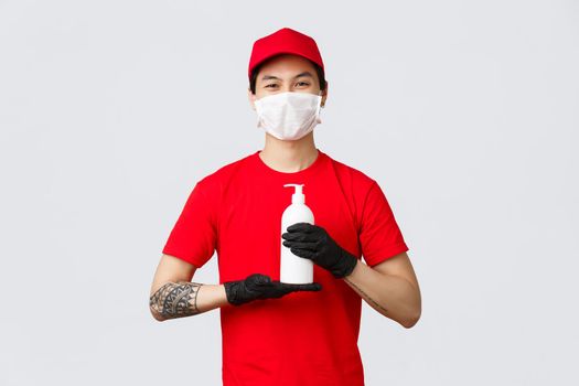 Safety contactless delivery, carriers concept. Smiling courier in medical mask, gloves and red uniform, holding hand sanitizer, suggest clients stay safe home while deliver parcels during covid 19.