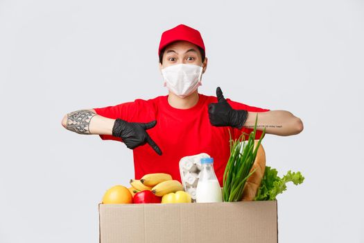 Online shopping, food delivery and coronavirus pandemic concept. Charismatic delivery guy in red uniform, medical mask and gloves, show thumb-up and approval, pointing at grocery parcel.