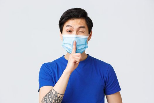 Different emotions, lifestyle and leisure during coronavirus, covid-19 concept. Cheeky handsome asian man in medical mask promise keep secret, shush and wink coquettish, be silent.