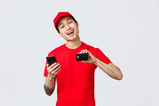 Friendly delivery guy in red uniform t-shirt and cap, showing smartphone screen and credit card, recommend application for tracking order online shopping. Courier advertise carrier service.