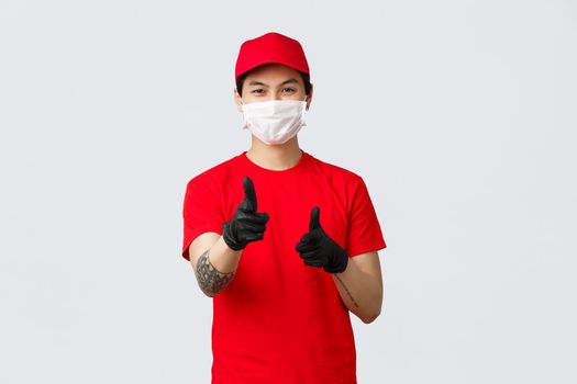 Happy delivery guy in medical mask and gloves, smiling with eyes as pointing camera, recommend carrier company service, provide fast and safe courier shipping, advertising and promoting.