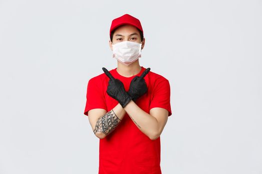 Young confident delivery guy in red t-shirt and cap, wear protective gloves and medical mask to deliver parcels at customer home during covid-19, showing variants, pointing sideways, two choices.
