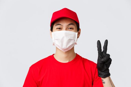 Covid-19, self-quarantine online shopping concept. Asian courier staying positive, friendly showing peace sign in medical mask and gloves, measures for safe order deliver on self-quarantine.