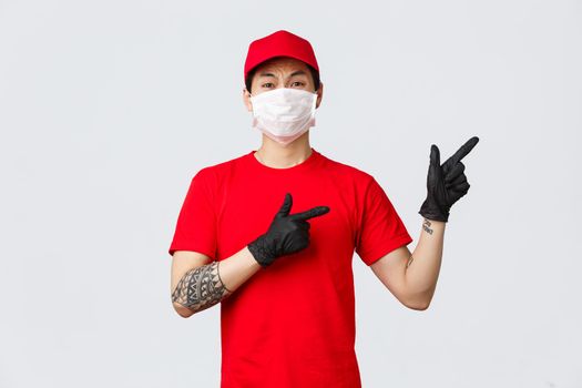 Anxious bothered asian delivery guy complaining over something, frowning and grimacing dislike, wear protective mask and medical gloves, pointing fingers right disappointed, showing bad thing.
