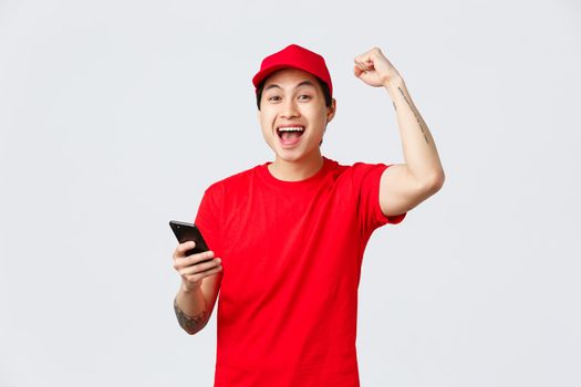 Happy, cheerful asian delivery guy in red uniform cap and t-shirt of carrier service, holding smartphone, reading good news, chanting with fist pump, yell yes celebrate victory or achivement.