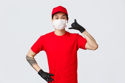 Cheerful asian delivery guy in medical mask and protective gloves show phone sign, asking clients give call anytime when delivery needed. Charismatic courier promote fast shipment of parcel.