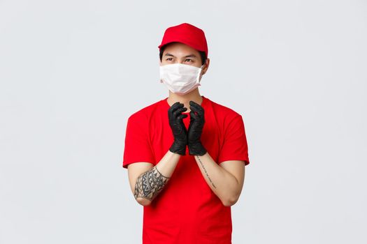 Creative asian delivery guy making plan, sheming something, have interesting thought in mind, steeple fingers in protective gloves, look away pondering idea, protect health with medical mask.