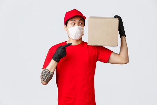 Concept of delivery and carriers during coronavirus pandemic. Intrigued delivery guy in uniform, courier service bring box to client, pointing and staring at package curiously, wear mask and gloves.