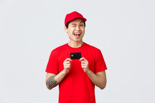 Friendly smiling asiand elivery man in red cap and t-shirt uniform, wink cheerful, recommend using credit card, buy contactless during virus outbreak. Courier give advice order online on coronavirus.