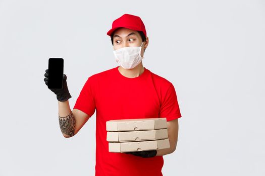 Funny delivery guy in red cap, t-shirt, holding pizza boxes and show smartphone screen. Asian courier confirm order info with client, bring food to people home during quarantine, wear medical mask.