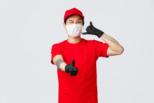 Call out customer support anytime. Cheerful asian delivery guy in medical mask and protective gloves, wink and show thumb-up, phone sign, provide safe courier service. carrier and shopping concept.