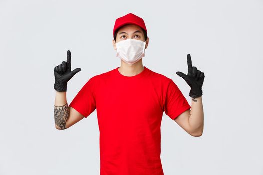 Intrigued cute asian delivery guy with tattoo, wearing uniform cap and red t-shirt, pointing fingers up, looking top advertisement with interest, reading sign upwards. Courier promote company service.