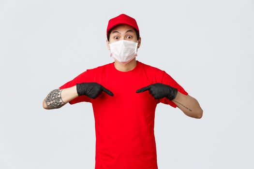 Surprised gasping asian delivery man in red cap and t-shirt, wearing protective mask and medical gloves to prevent virus spread, care for customers safety during self-qurantine online orders delivery.