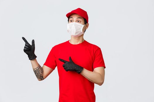 Impressed, shocked asian delivery man in medical mask and protective gloves, seeing something exciting, pointing looking upper left corner. Courier show advertisement, wearing company uniform.