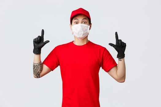 Surprised young asian delivery guy in company uniform, red t-shirt and cap, staring camera impressed and shocked, pointing fingers up, wear protective gloves and medical mask while at work.