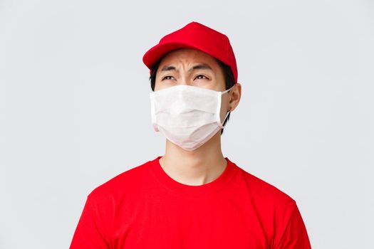 Covid-19, self-quarantine online shopping and delivery concept. Complaining reluctant courier in red t-shirt and cap, medical mask, whining about night shifts at work, look disappointed.