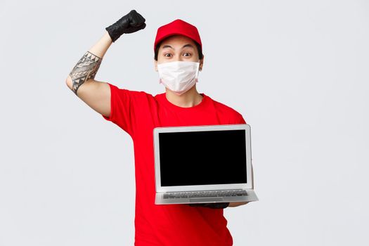Cheerful happy asian delivery guy deliver parcels during covid 19 pandemic, safe shopping online. Courier holding laptop and showing advertisement on screen, chanting, raise hand in celebration.