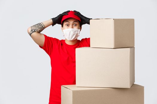 Nervous asian delivery guy got lots of orders to deliver, grab head in panic, standing near piles of boxes. Courier in medical mask and gloves feeling anxious working, clients waiting for packages.