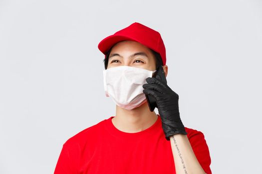 Covid-19, self-quarantine online shopping and delivery concept. Friendly young asian courier in medical mask and protective gloves, calling client, answer customer phonecall, smiling, grey background.