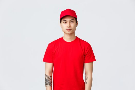 Express delivery, shipping and logistics concept. Young handsome asian courier in red cap and t-shirt, looking camera, waiting for client order delivered company storage.