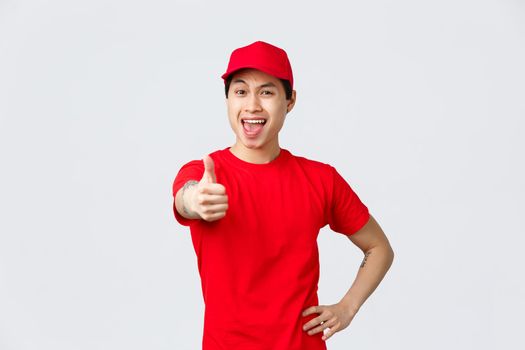 Cheerful delivery guy in uniform encourage make more orders shopping online. Handsome asian courier thumb-up, guarantee best quality or parcel shipping, transfer packages to clients.