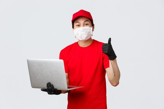 Happy asian delivery man in medical mask, gloves show thumb-up in approval, recommendation. Courier holding laptop, received new order for delivery, confirm shipping, customer support answer client.