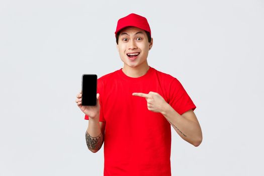 Delivery application, online shopping and shipping concept. Surprised asian courier in red cap and t-shirt, pointing finger at mobile phone app, showing smartphone screen with amused face.