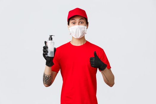 Amused delivery man in red cap and t-shirt, make thumb-up sign, wear protective gloves and medical mask, provide clients and company workers with hand sanitizers. Concept of contactless safe delivery.