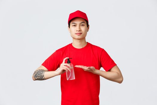 Quarantine, safe contactless delivery and online shopping concept. Smiling asian delivery guy in red cap and t-shirt, apply hand sanitizer on hands to prevent spread of covid-19 and viruses.