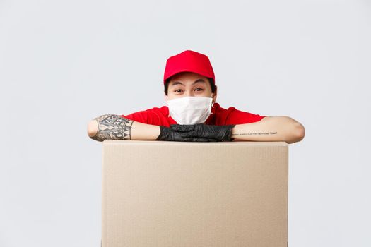 Delivery, online shopping and quarantine concept. Cheerful asian delivery guy in red cap uniform, medical mask and protective gloves, leaning on large box, order or parcel, promote courier service.