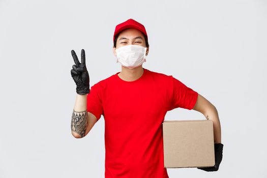 Concept of delivery, coronavirus pandemic. Cheerful smiling asian courier in red cap, t-shirt, show peace sign, holding parcel, wear medical mask and gloves for client safety, deliver package.