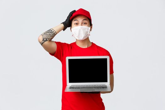 Excited, happy delivery guy happy, touching head from surprise, showing carrier webpage laptop screen, wear protective medical mask and gloves, courier uniform, shipping of package during pandemic.