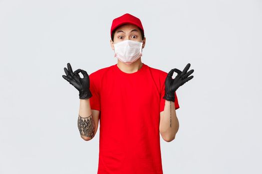 Worried young asian delivery man trying calm down nervous client, showing zen gesture, wearing medical mask and protective gloves, company uniform. Courier make okay sign ensure best quality.